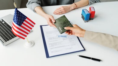 A person hands a passport holder across the table to another person. On the table is an approved form and a U.S. Flag.