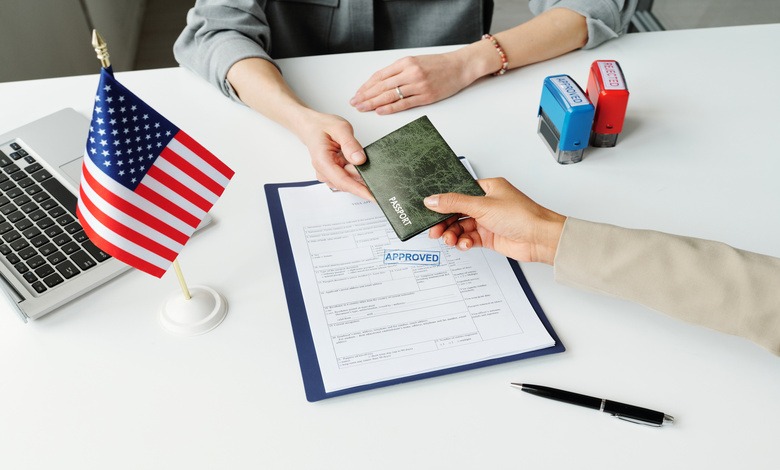 A person hands a passport holder across the table to another person. On the table is an approved form and a U.S. Flag.