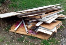 A large pile of scrap wood lies on patchy grass. Some of the wood is brown while other pieces are white.