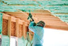 A man wearing a blue protective gear and mask applies spray foam insulation to the roof frame of a wooden house.