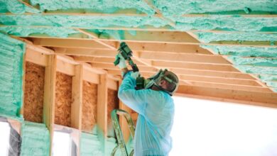 A man wearing a blue protective gear and mask applies spray foam insulation to the roof frame of a wooden house.