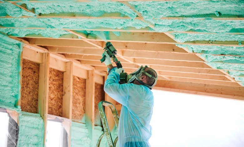 A man wearing a blue protective gear and mask applies spray foam insulation to the roof frame of a wooden house.