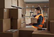 A warehouse stocker holding a box in her hand and scanning the bar code with a scanner. Various boxes surround her.