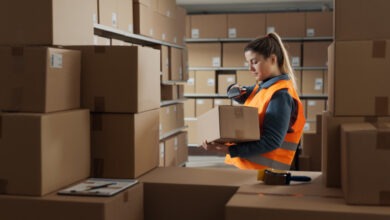 A warehouse stocker holding a box in her hand and scanning the bar code with a scanner. Various boxes surround her.