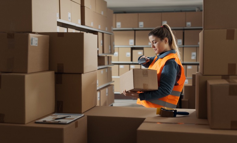 A warehouse stocker holding a box in her hand and scanning the bar code with a scanner. Various boxes surround her.