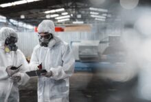 Two workers wearing full-body hazmat suits and full-face respirator masks stand in a warehouse, reviewing a document.