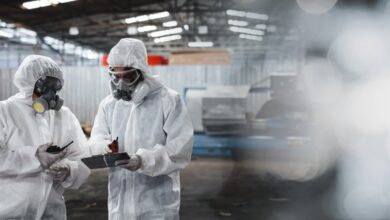 Two workers wearing full-body hazmat suits and full-face respirator masks stand in a warehouse, reviewing a document.
