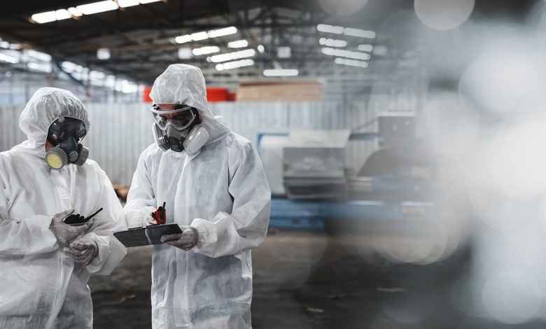 Two workers wearing full-body hazmat suits and full-face respirator masks stand in a warehouse, reviewing a document.