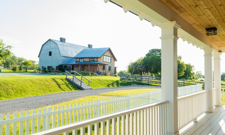 The view from a porch with white railings. A barn-style house with a gravel driveway sits in the distance.