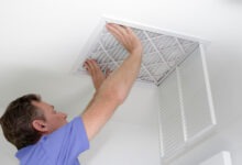 A brown-haired man in a blue shirt uses two hands to push a clean air filter into a white ceiling vent.