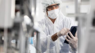 A man wearing a hairnet, goggles, gloves, and a white robe, holding a small tablet in between two blurred machines.