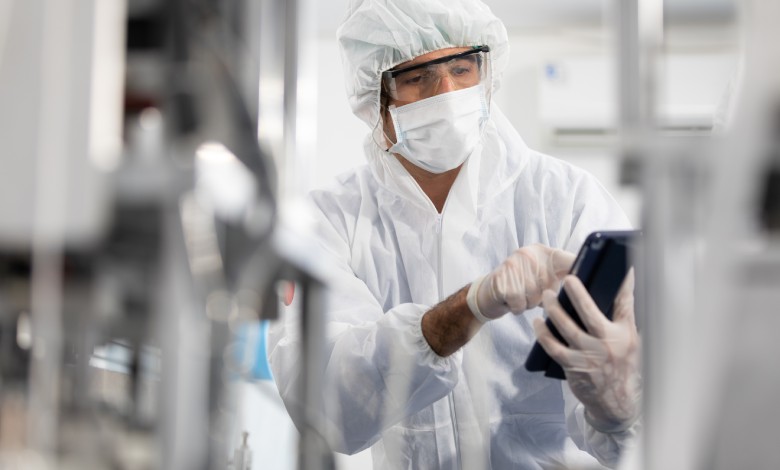 A man wearing a hairnet, goggles, gloves, and a white robe, holding a small tablet in between two blurred machines.