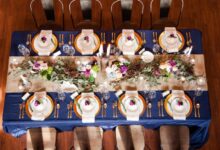 An overhead angle of a rectangular table. The table has a navy tablecloth, a beige runner, and gold chargers.