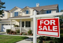 A for sale sign in front of a two-story house with a porch. The lawn, with flowers by the porch, is well-maintained.