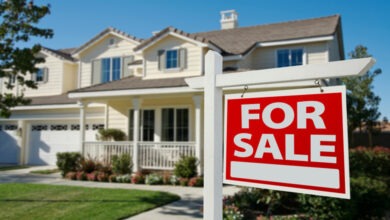 A for sale sign in front of a two-story house with a porch. The lawn, with flowers by the porch, is well-maintained.