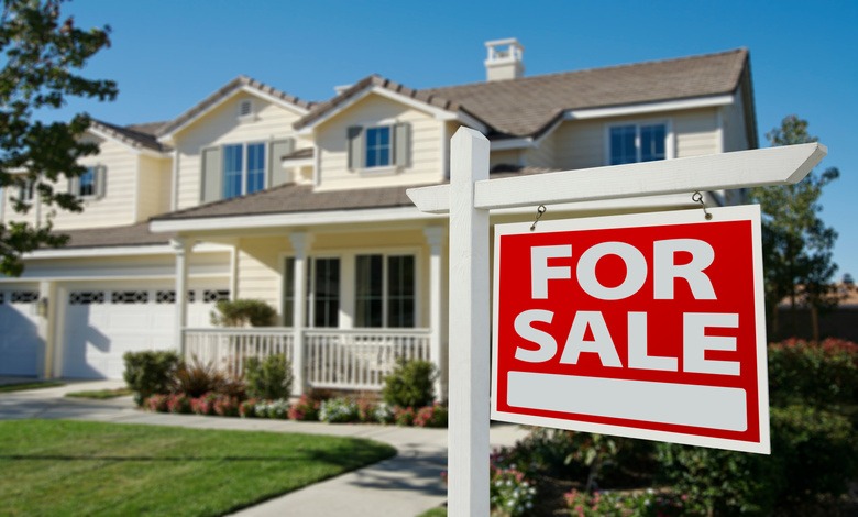A for sale sign in front of a two-story house with a porch. The lawn, with flowers by the porch, is well-maintained.