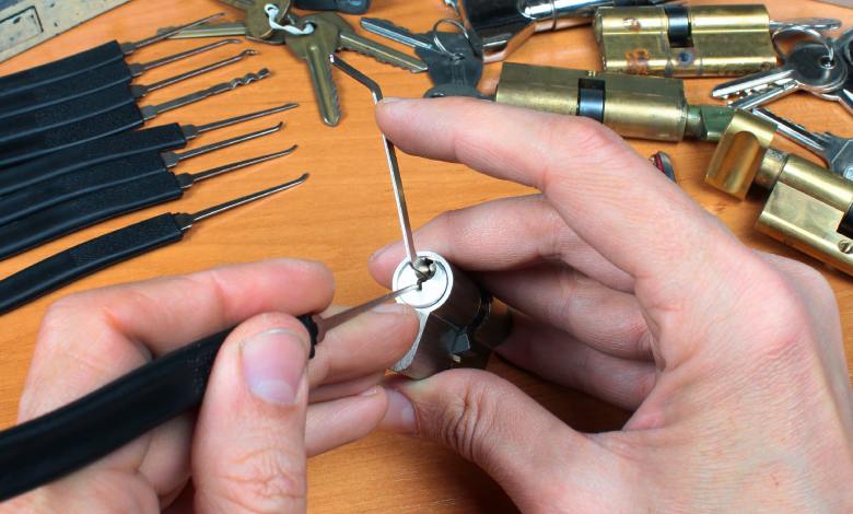 Close-up of a pair of hands trying to pick a cylinder lock with a lockpick and a tension wrench. Other tools lie nearby.