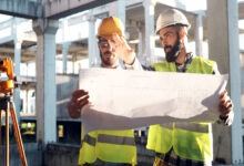 Two male engineers wearing hard hats and safety vests stand side by side while holding a blueprint of the site.