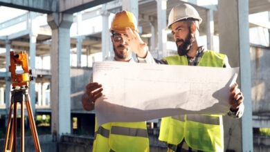 Two male engineers wearing hard hats and safety vests stand side by side while holding a blueprint of the site.