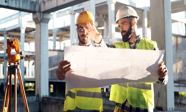 Two male engineers wearing hard hats and safety vests stand side by side while holding a blueprint of the site.