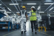 Two people are walking side by side in a room full of heavy machinery. They’re both wearing hard hats.