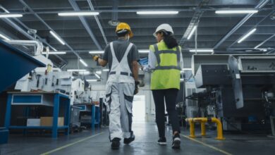 Two people are walking side by side in a room full of heavy machinery. They’re both wearing hard hats.