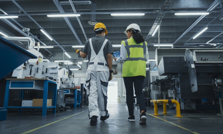 Two people are walking side by side in a room full of heavy machinery. They’re both wearing hard hats.