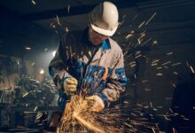 A man wearing a helmet, safety gloves, and a uniform with thick fabric, using a grinder producing multiple sparks.