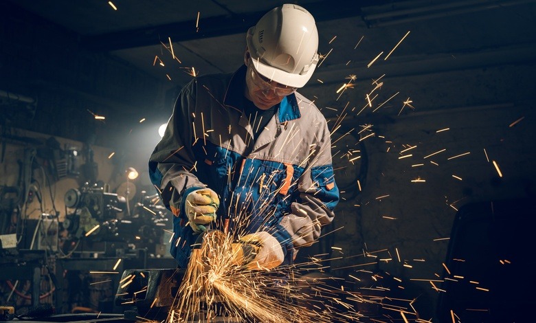 A man wearing a helmet, safety gloves, and a uniform with thick fabric, using a grinder producing multiple sparks.