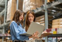 Two women stand next to a large shelf in a warehouse. Both look at a laptop that one of the women holds in her arms.