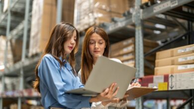 Two women stand next to a large shelf in a warehouse. Both look at a laptop that one of the women holds in her arms.
