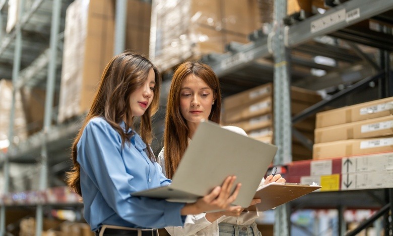 Two women stand next to a large shelf in a warehouse. Both look at a laptop that one of the women holds in her arms.