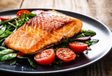 A large piece of salmon with mixed vegetables, including spinach and tomatoes, on a round black plate.