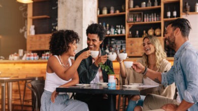 Two men and two women sit at a square table in a coffee shop, smiling and laughing while holding white mugs.