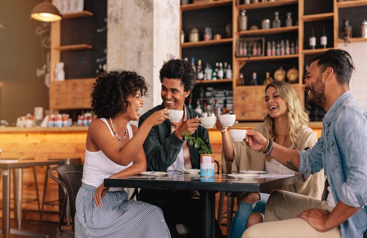 Two men and two women sit at a square table in a coffee shop, smiling and laughing while holding white mugs.