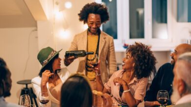 A group of seven people at a nighttime get-together in a warmly lit home. The host pours wine for a seated guest at a table.