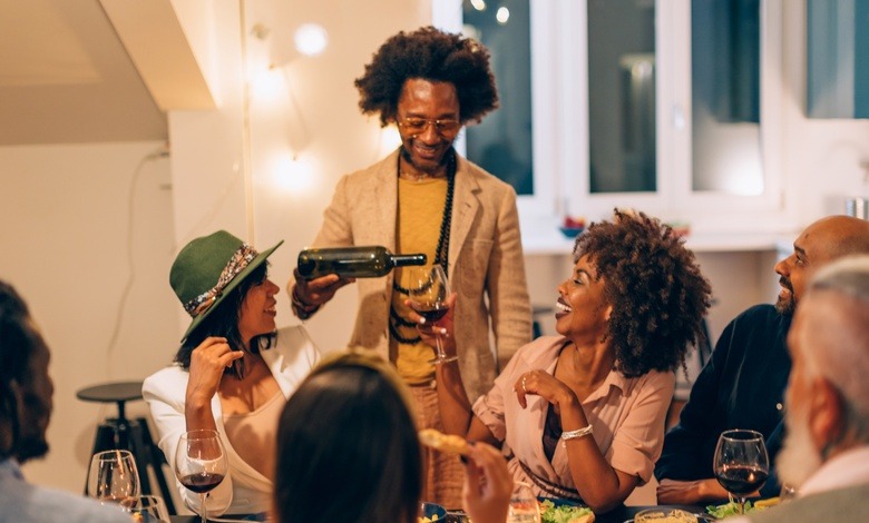 A group of seven people at a nighttime get-together in a warmly lit home. The host pours wine for a seated guest at a table.