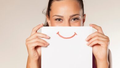 A woman holds a piece of paper over her face that has a smile printed on it. She stands in front of a white background.