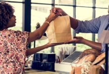 A man wearing an apron hands a woman a brown bag over a checkout counter. Loaves of bread are on the counter.