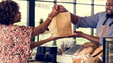 A man wearing an apron hands a woman a brown bag over a checkout counter. Loaves of bread are on the counter.
