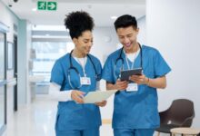 Two nursing professionals in blue scrubs review patient information on a tablet and paper in a modern hospital hallway.