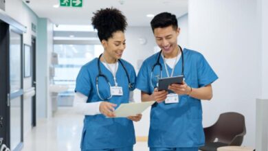 Two nursing professionals in blue scrubs review patient information on a tablet and paper in a modern hospital hallway.