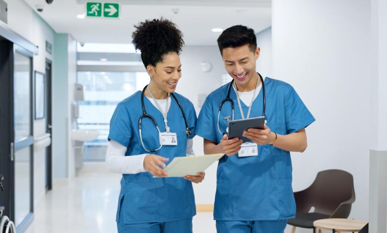 Two nursing professionals in blue scrubs review patient information on a tablet and paper in a modern hospital hallway.