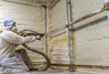 Worker in full protective gear sprays foam insulation onto wall studs inside a building under construction.