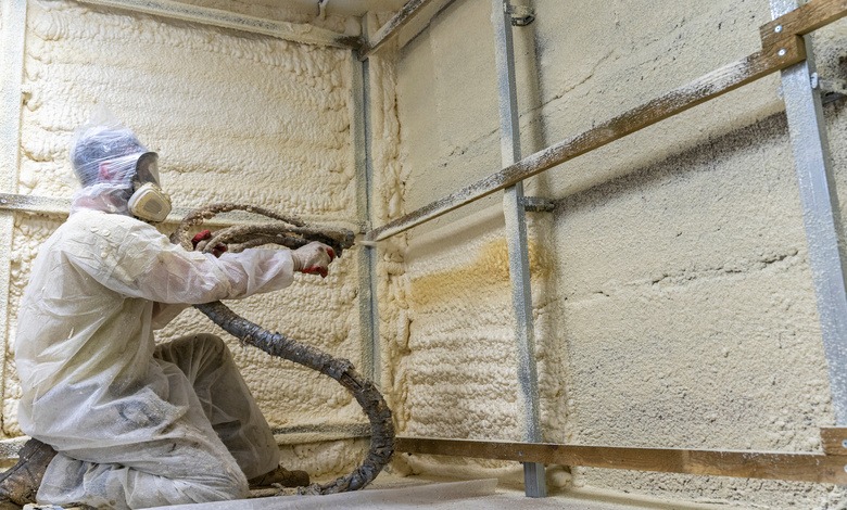 Worker in full protective gear sprays foam insulation onto wall studs inside a building under construction.