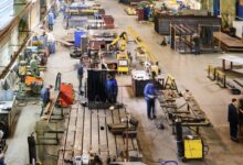A large manufacturing shop floor with several metalworking machines, workbenches, and workers welding and assembling components.