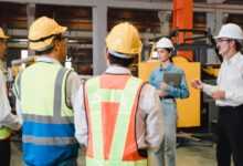 A group of four warehouse workers wearing hard hats and protective vests stand across from two inspectors wearing business casual clothing.