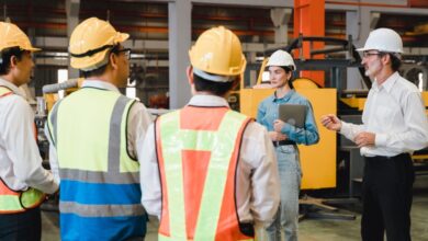 A group of four warehouse workers wearing hard hats and protective vests stand across from two inspectors wearing business casual clothing.