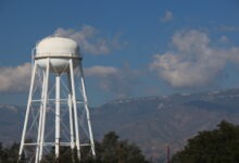 A tall white water tower with a spherical tank in front of mountains. The tower is framed by trees and utility poles.