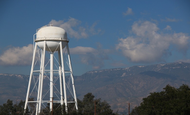 A tall white water tower with a spherical tank in front of mountains. The tower is framed by trees and utility poles.